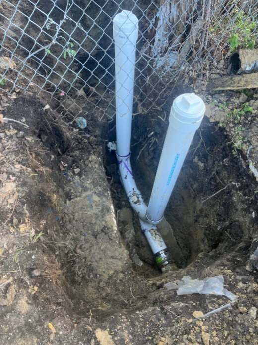 Exposed PVC commercial drain lines being repaired near a chain link fence